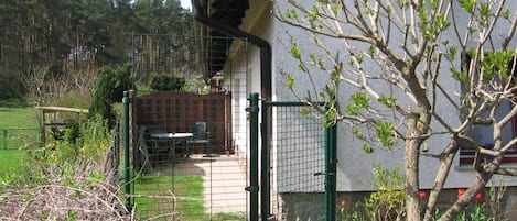 A fenced outdoor area with a table and chairs, a building with a red roof, and a tree with green leaves.