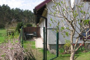 Ein von einer Hecke umgebener Außenbereich mit Tisch und Stühlen, ein Gebäude mit rotem Dach und ein Baum mit grünen Blättern.