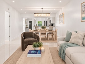 A modern living room with a sofa, a coffee table with a book and a plant, and a dining area with a table set for two. - Renovated PGA National Home by Guaranteed Rental (Palm Beach Gardens)