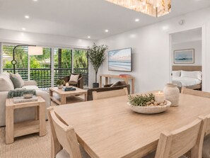 A modern living room with a wooden dining table, a sofa, and a coffee table. There is a large window with a view of greenery outside. - Renovated PGA National Home by Guaranteed Rental (Palm Beach Gardens)