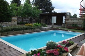 A rectangular swimming pool with a wooden deck surrounding it, a stone wall with a wooden pergola, and various potted plants. - Holiday apartment with 1 bedroom (Dresden OT Borthen)