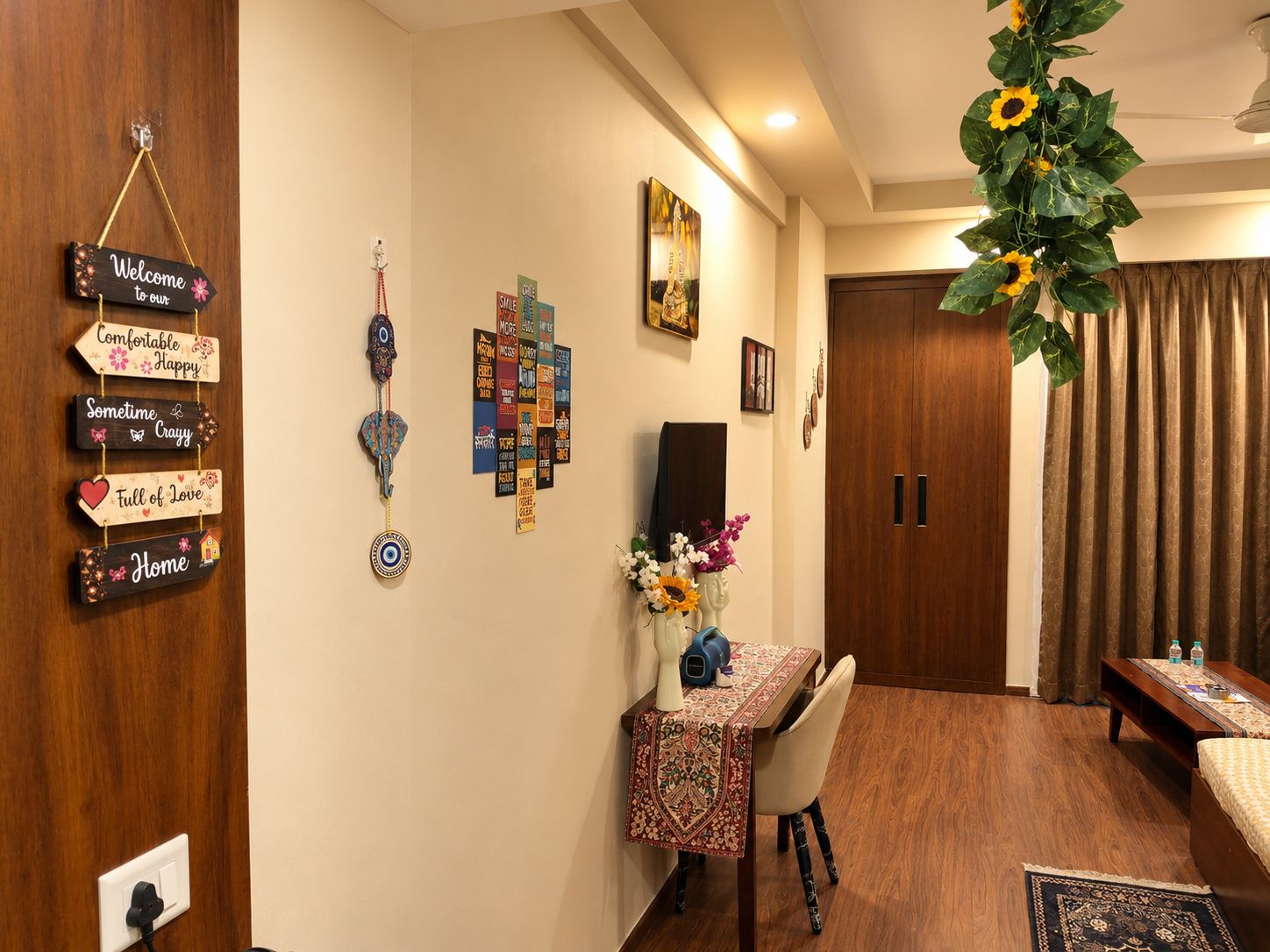 A cozy hotel room with a wooden door, a desk with a patterned tablecloth, a chair, a bed, and a hanging plant with sunflowers.