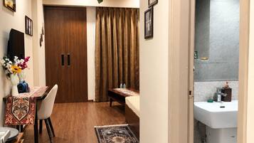 A hotel room with a wooden floor, a dining area with a table and chairs, a bed, a bathroom with a sink and mirror, and a hanging plant.
