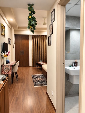 A hotel room with a wooden floor, a dining area with a table and chairs, a bed, a bathroom with a sink and mirror, and a hanging plant.