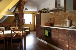 A kitchen with wooden cabinets, a dining table with chairs, a hanging light fixture, and a window with curtains. - Holiday apartment with 1 bedroom (Lübben \/ Spreewald)