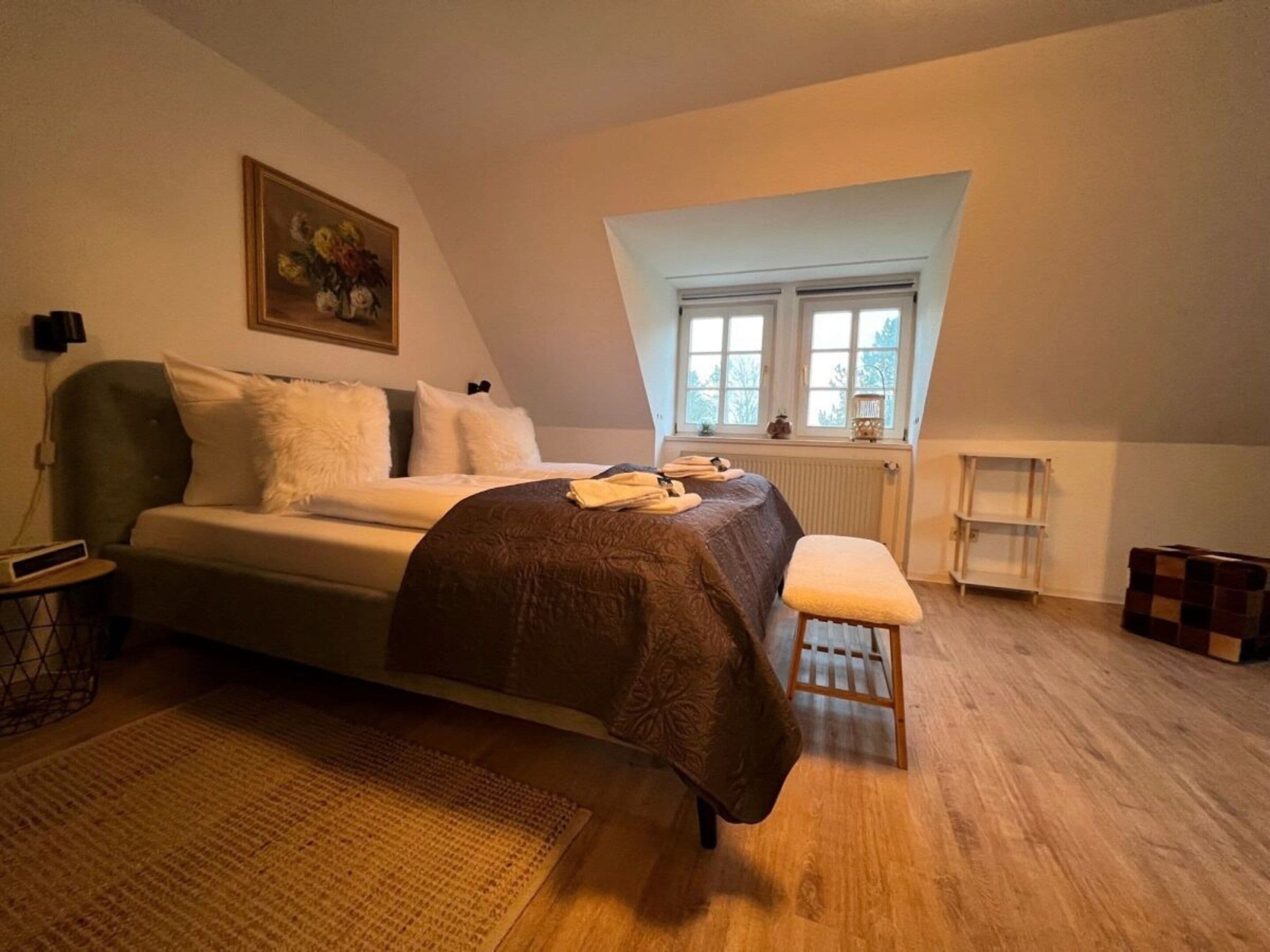 A bedroom with a bed, a bench, a radiator, a window, and a painting on the wall.