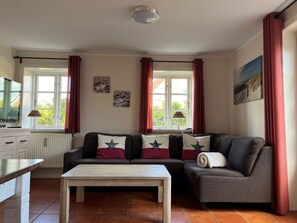 Ein Wohnzimmer mit einer Couch, einem Couchtisch und einem Fernseher. Es gibt Fenster mit Vorhängen, eine Heizung und Kunstwerke an den Wänden.