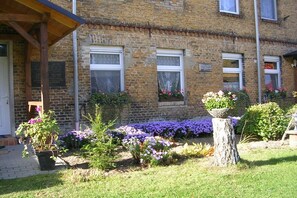 Un edificio de ladrillo con un porche cubierto, ventanas con jardineras y un jardín con diversas plantas y flores.