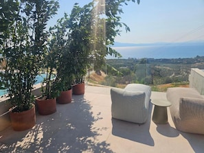 A terrace with potted plants, outdoor furniture, and a view of the sea and landscape.
