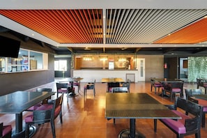 Interno moderno di un ristorante con zona pranzo centrale, cucina sullo sfondo e area bar con schermo televisivo.