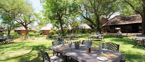 An outdoor dining area with tables and chairs, surrounded by trees and buildings with thatched roofs.