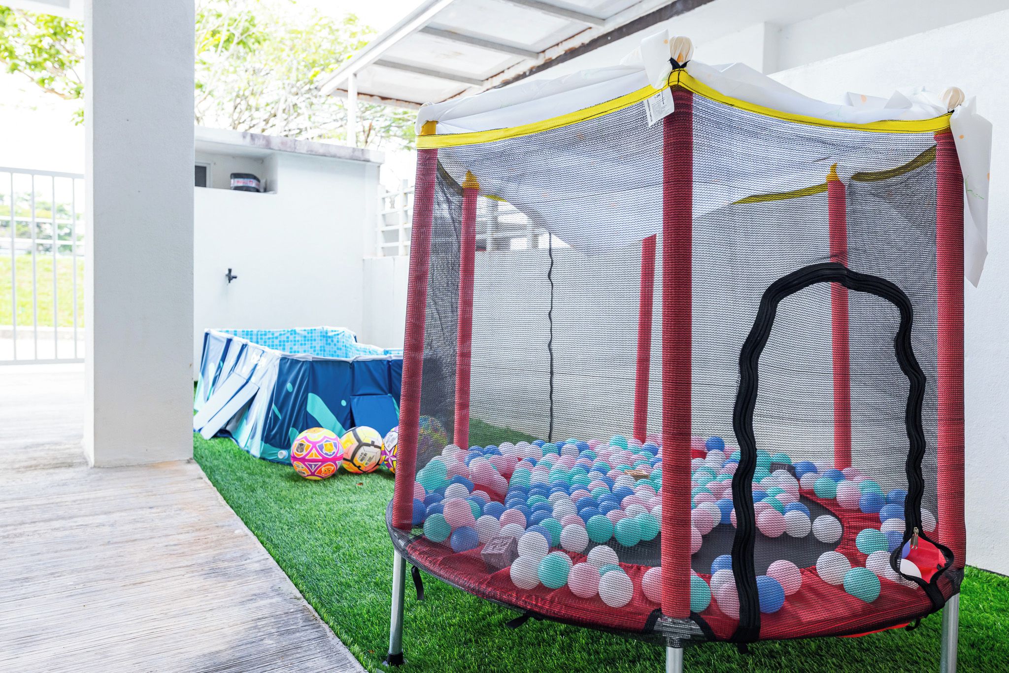 A trampoline with a safety net enclosure, surrounded by colorful balls, under a covered area with artificial grass.