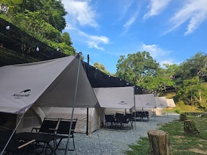 Una fila de tiendas con la marca 'Natureline', dispuestas sobre un sendero de grava con sillas al frente, rodeadas de árboles.