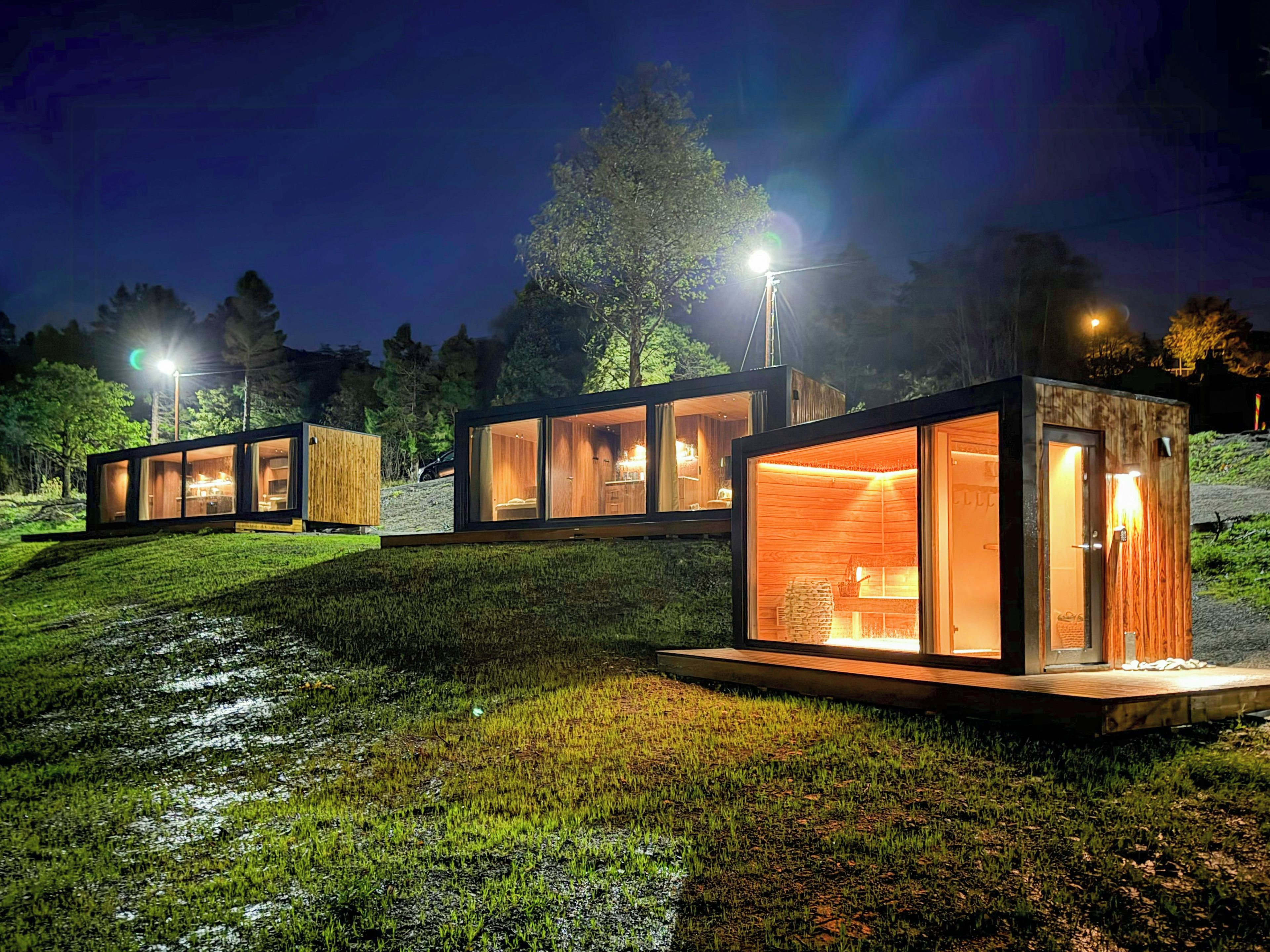Estrutura moderna com paredes de vidro e exterior de madeira, localizada em um declive gramado com o céu noturno ao fundo.