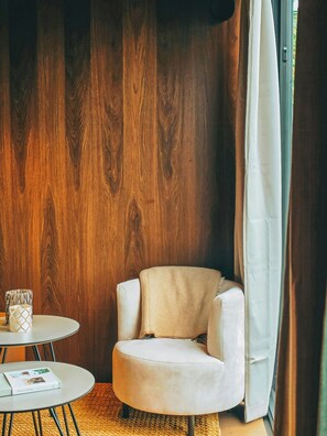 Quarto com parede de madeira, uma poltrona branca, uma pequena mesa redonda com um livro e uma janela com cortinas.