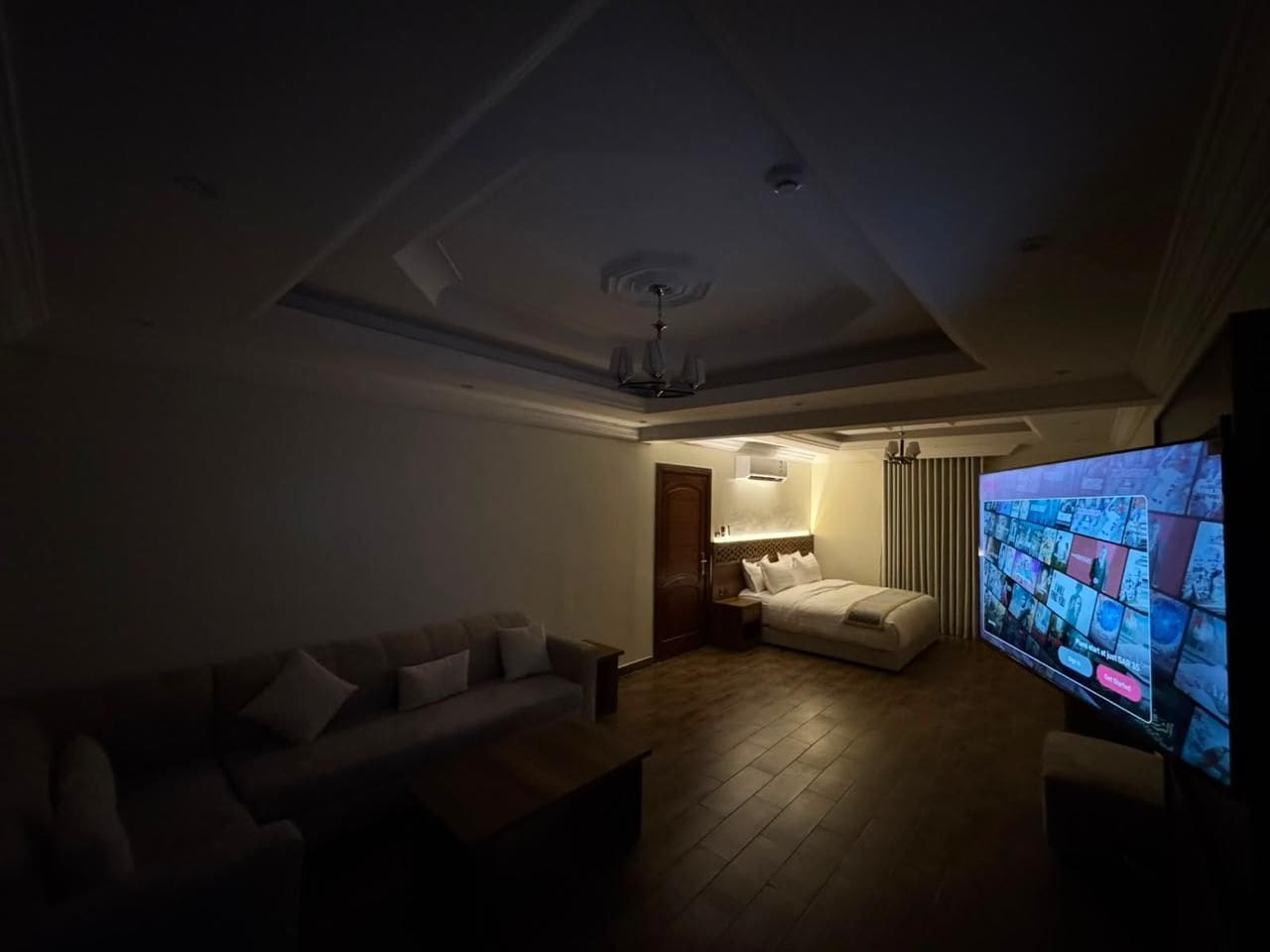 A dimly lit living room with a sofa, a bed, and a television displaying a news channel.