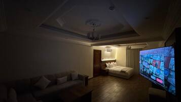 A dimly lit living room with a sofa, a bed, and a television displaying a news channel.
