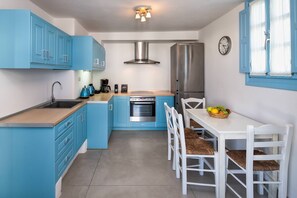 A compact kitchen with blue cabinets, a stainless steel refrigerator, a built-in oven, and a dining area with white chairs and a fruit basket. - My Rental Homes - Guest House Skiron, with private pool and sea view (Fira)
