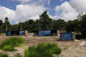 Uma cabana moderna e minimalista com grandes janelas de vidro, localizada em um ambiente natural com árvores e céu claro.