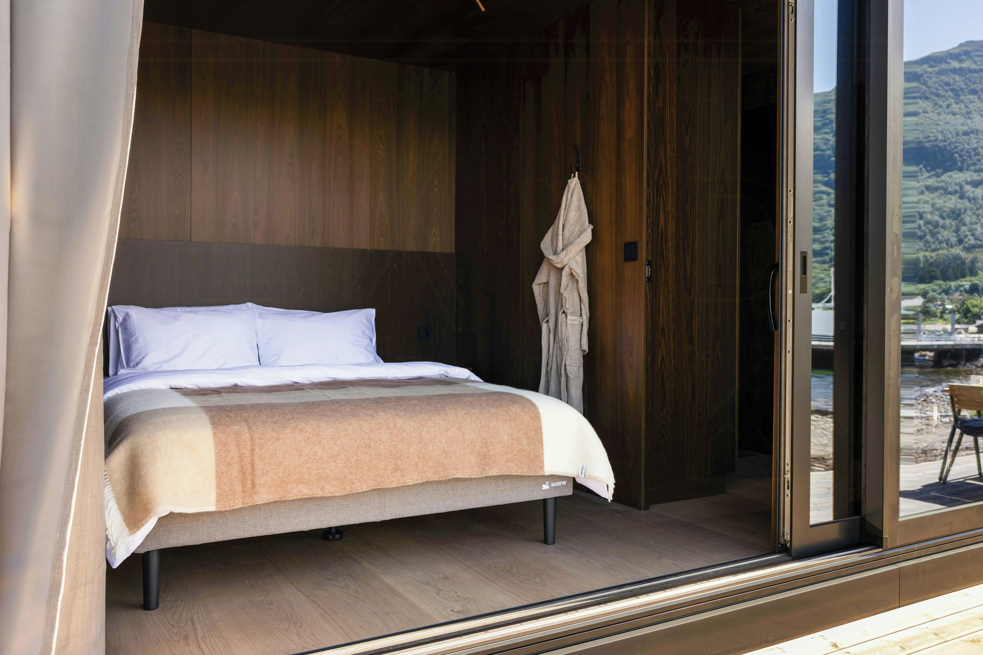 Quarto com cama, paredes de madeira e vista para uma paisagem através de uma porta de vidro.