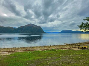 Um lago com montanhas ao fundo, uma área gramada em primeiro plano e um banco.