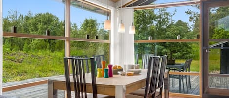 An open-plan dining area with a wooden table and chairs, large windows, and a view of greenery.