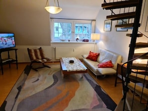 A living room with a sofa, a patterned rug, a television, and a window with a view of a house. - Holiday Home in Nieblum With Swimming Pool (Nieblum)
