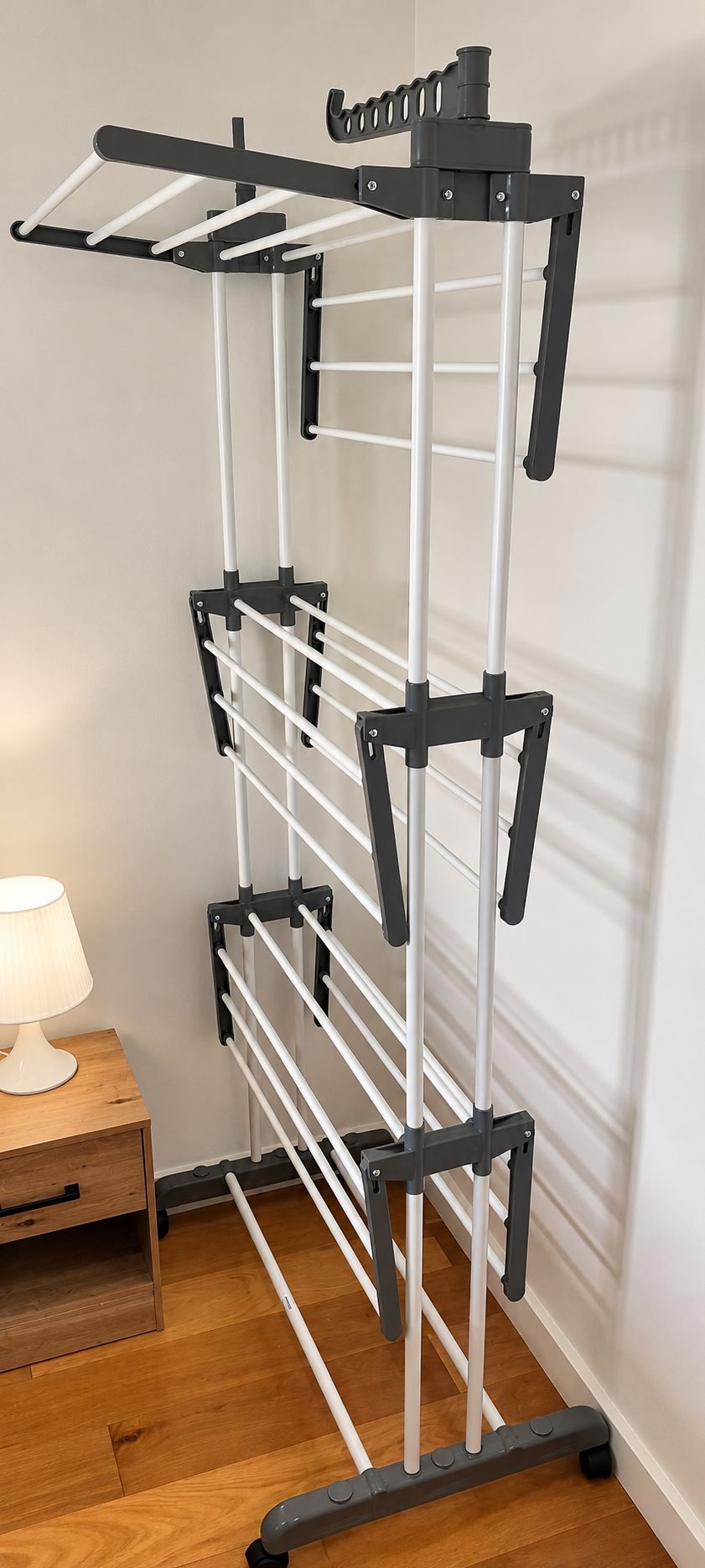 A wall-mounted drying rack with multiple pegs, a wooden nightstand with a lamp, and a wooden floor.