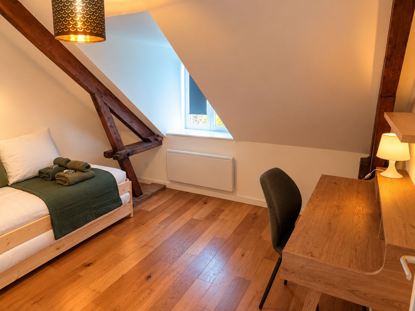 A bedroom with a sloped ceiling, a bed with a green blanket, a wooden desk, and a chair.