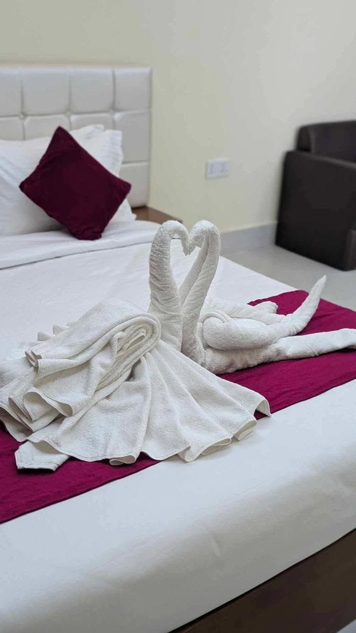 A neatly made bed with a heart-shaped towel arrangement on a white bedspread and a magenta throw.