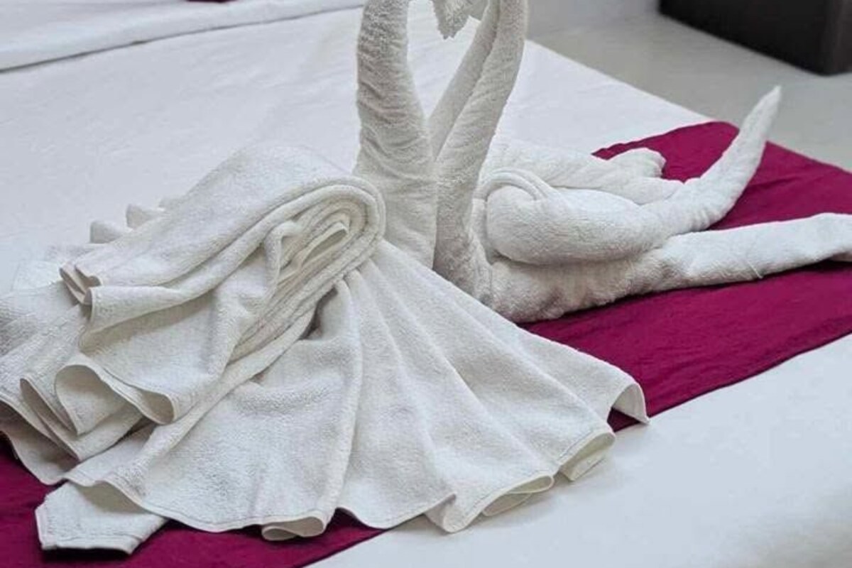 Un lit bien fait, agrémenté d’une serviette pliée en forme de cœur sur un couvre-lit blanc et d’un plaid magenta.