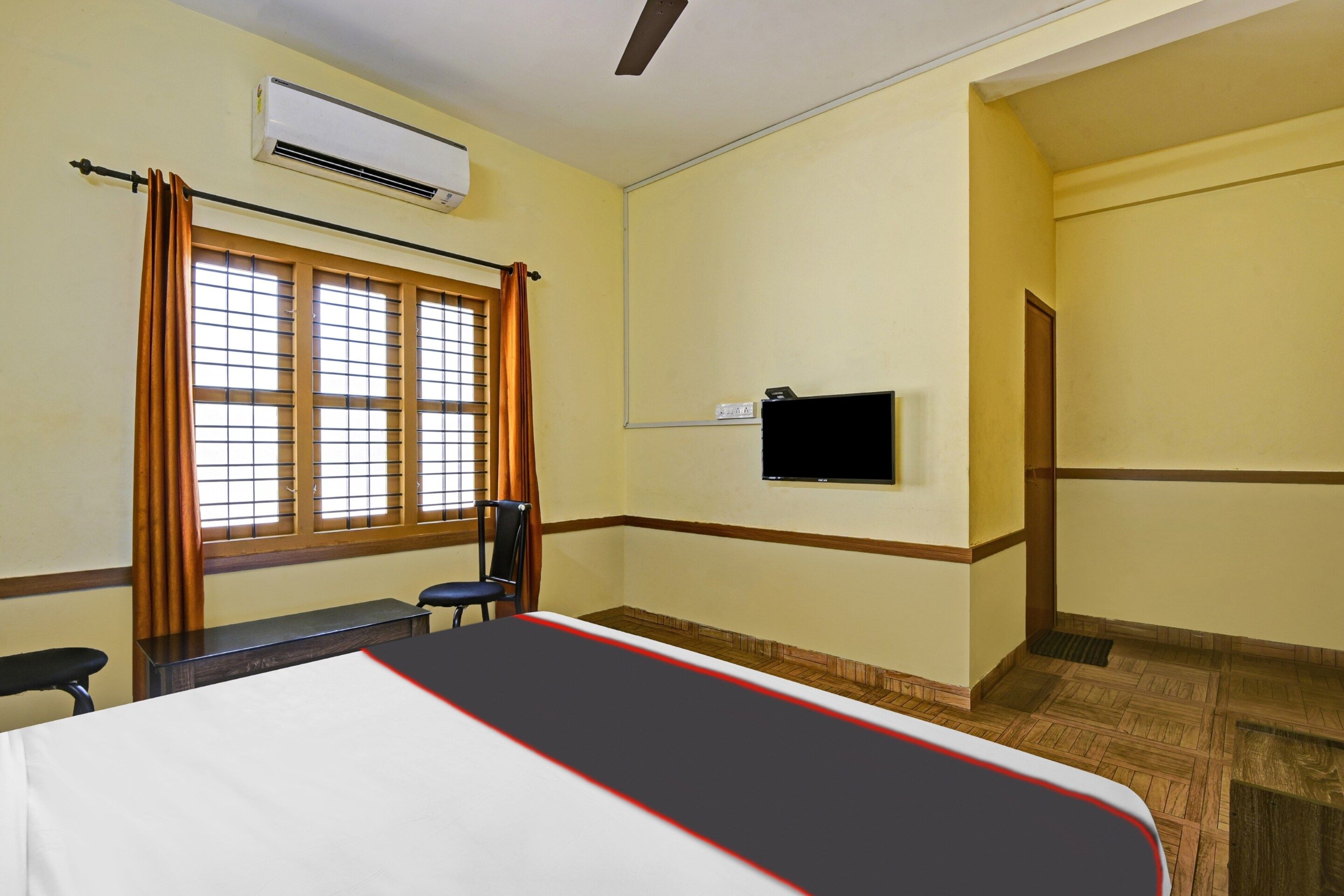Una habitación de hotel con cama, una silla, un equipo de aire acondicionado, televisor y ventana con cortinas.