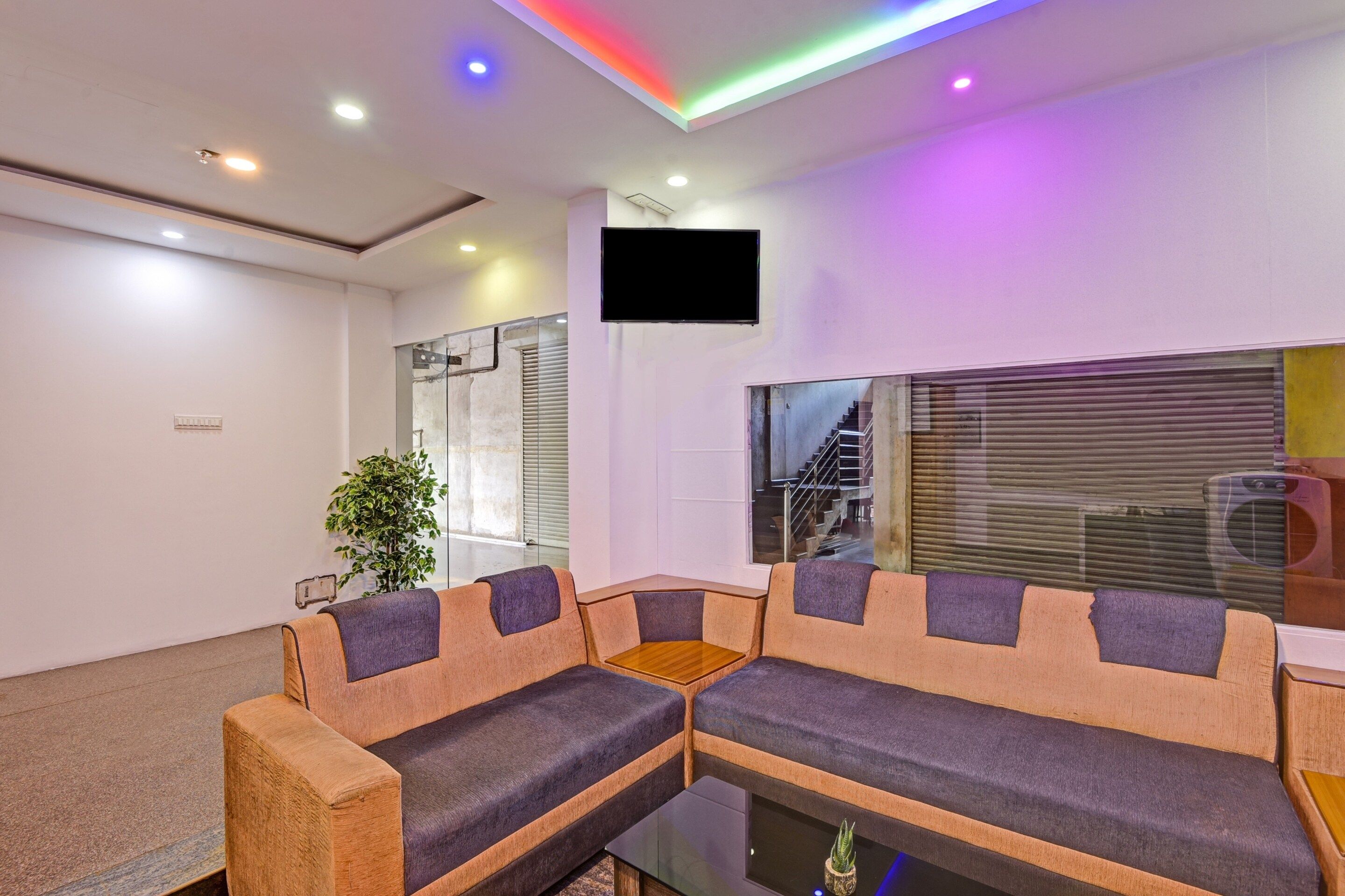 A modern living room with a sectional sofa, a flat-screen TV mounted on the wall, and a staircase visible through a large window.