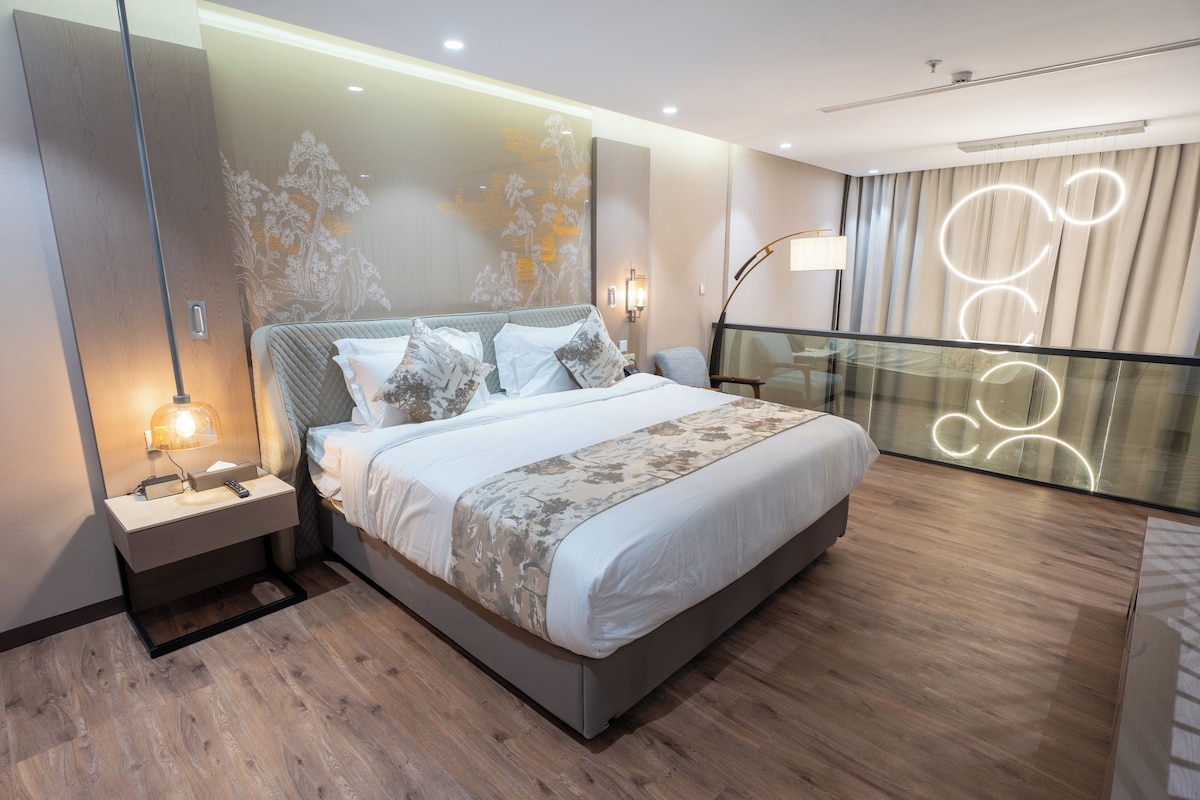 Une chambre d’hôtel moderne avec un grand lit, des tables de chevet, une petite table, une lampe et une vue sur la ville par la fenêtre.
