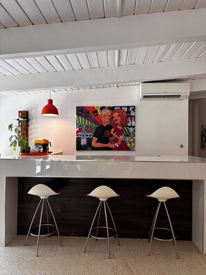 A modern kitchen with a white countertop, a red pendant light, a colorful abstract painting, and three white bar stools. - Mid-century designer home with pool and grill in plantation (Plantation)