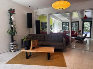 A modern living room with a sofa, a coffee table, and a bookshelf. There is a large window providing a view of the outdoors. - Mid-century designer home with pool and grill in plantation (Plantation)