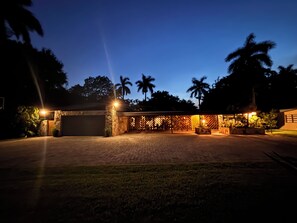 Un camino de entrada con portón que conduce a un edificio con un patio techado, palmeras y una entrada bien iluminada.