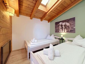 Een kamer met twee bedden, een dakraam, een schilderij van lammertjes en een klein tafeltje met een lamp.
