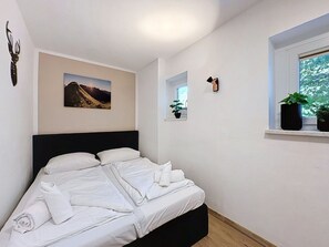 Ein ordentlich bezogenes Bett mit weißen Leinen, ein Gemälde mit Berglandschaft darüber, ein kleines Fenster mit einer Pflanze und eine wandmontierte Leuchte.