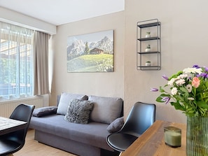 Ein modernes Wohnzimmer mit einem grauen Sofa, schwarzen Sesseln, einem Holztisch, einer Vase mit Blumen, einem gerahmten Bild an der Wand und einem schwarzen Metallregal mit Pflanzen.