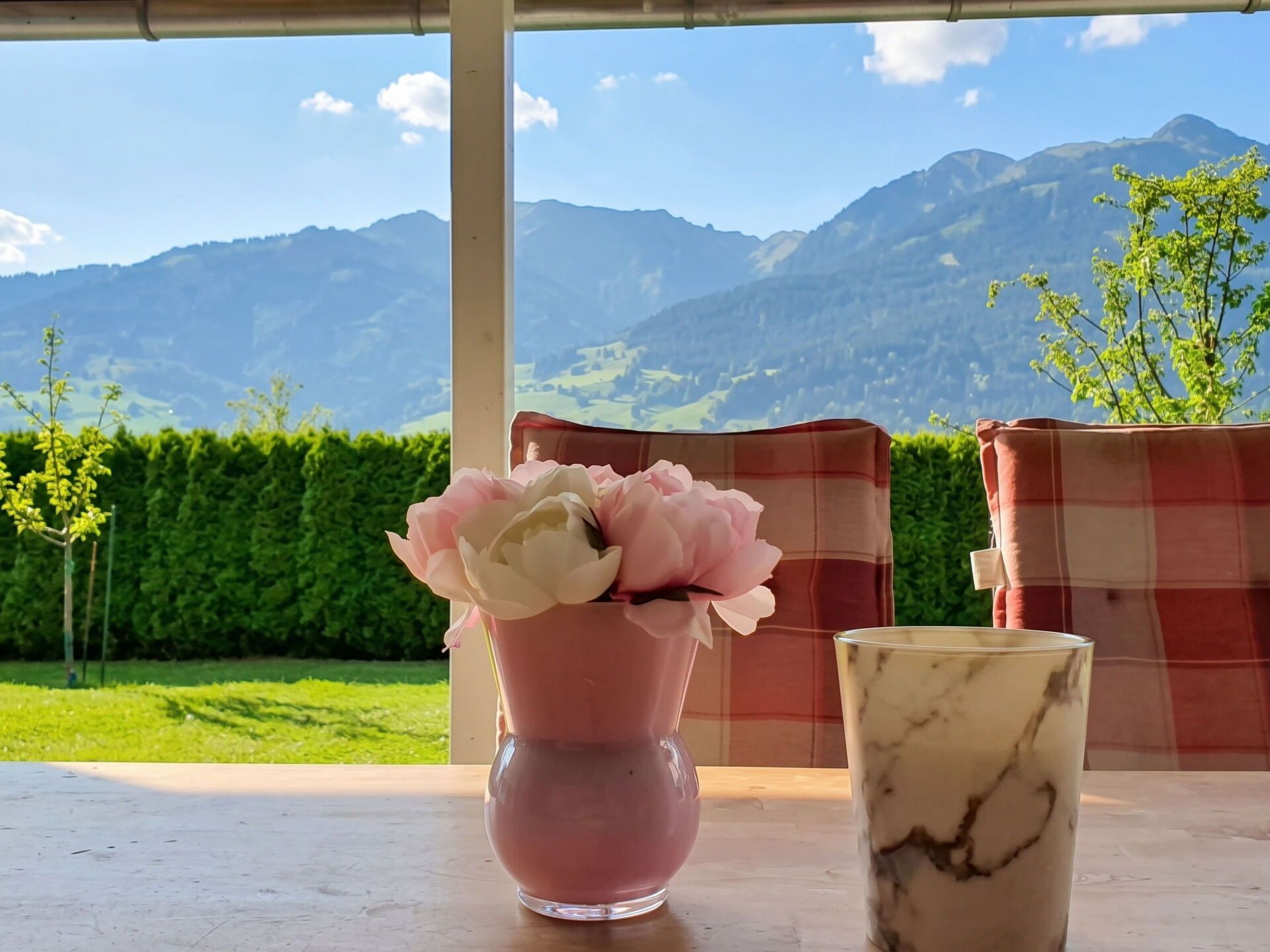 A table with a vase of flowers and a glass, set against a backdrop of mountains and greenery.