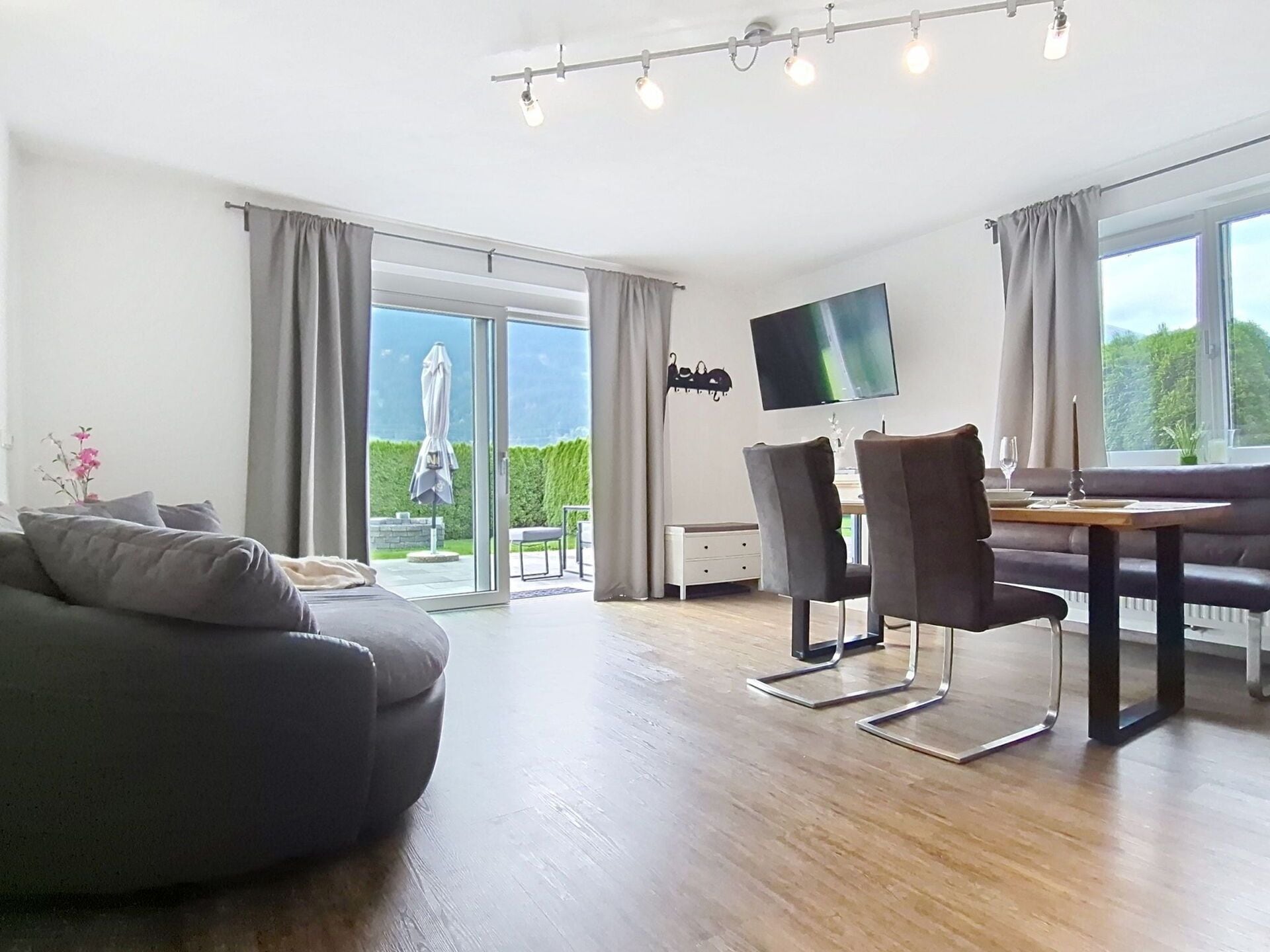 A modern living room with a sofa, dining table, chairs, and a flat-screen TV mounted on the wall.