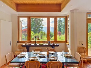 Comedor con mesa dispuesta para cuatro, ventanas amplias con vistas a árboles y montañas, y techo de madera.