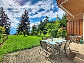 A patio with a table set for dining, surrounded by a lush garden and tall trees. - Nice apartment for 6 people with WIFI, TV, terrace, pets allowed and panoramic view (Crans-montana)