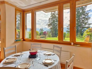 Ein Essbereich mit einem Tisch für vier Personen, großen Fenstern mit Holzrahmen und Blick auf einen Garten und Berge.