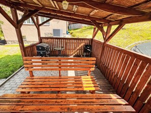 A wooden bench under a pergola with a grill and table.