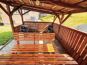 A wooden bench under a pergola with a grill and table.