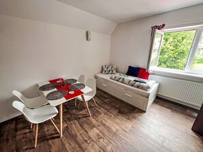 Ein gemütlicher Raum mit Esstisch, Stühlen, einer Bank mit Kissen, einem Heizkörper und einem Fenster mit Blick auf Grünflächen.
