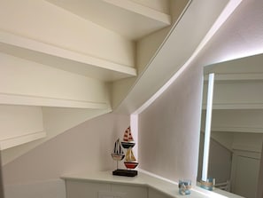 Un escalier avec une rampe blanche, une petite étagère ornée d’objets décoratifs et un grand miroir qui reflète l’intérieur.