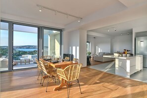 Eine geräumige, moderne Küche mit einer zentralen Kochinsel, einem Essbereich mit Holztisch und Korbstühlen sowie einer großen Glastür, die auf eine Terrasse mit Meerblick führt.